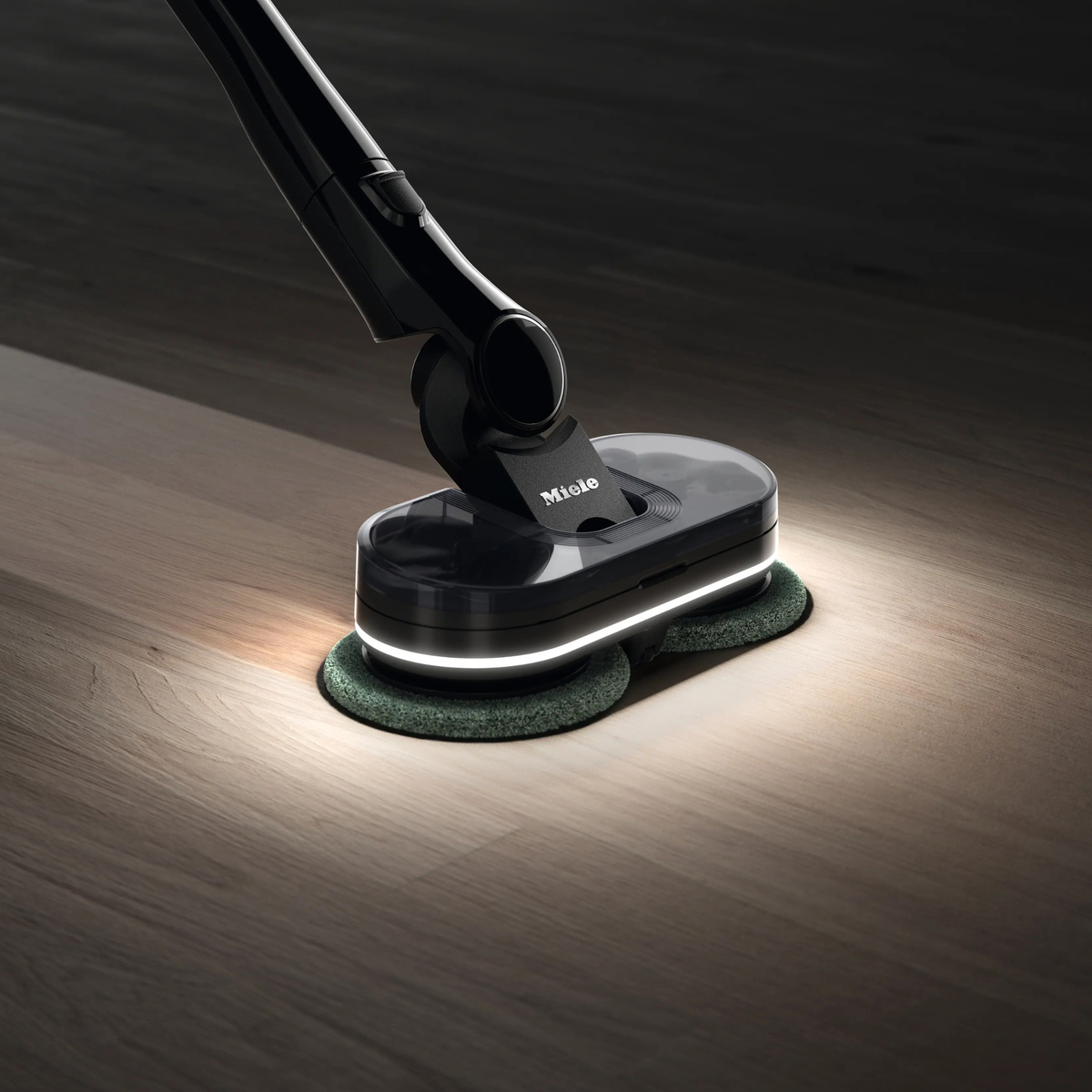 Miele AquaTwister wet-cleaning a dark hardwood floor, its SpotLight illumination revealing the cleaned path.