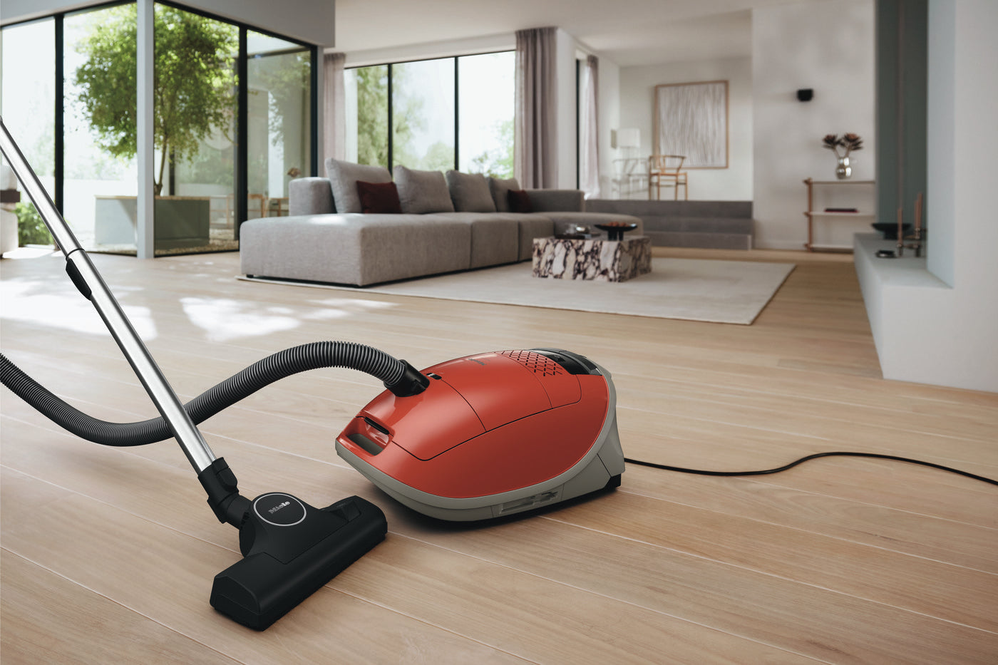 Red Miele Guard M1 Terra Red canister vacuum in a modern living room with wood floors.