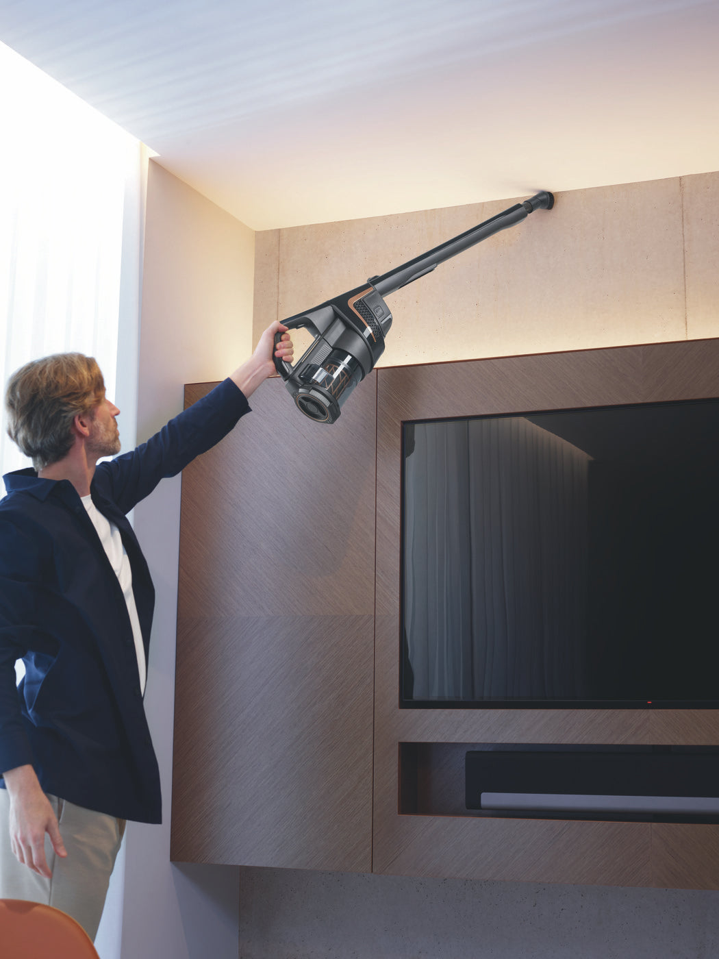 Woman using the Miele Triflex HX2 Cat & Dog's handheld unit to dust near a wall-mounted TV.