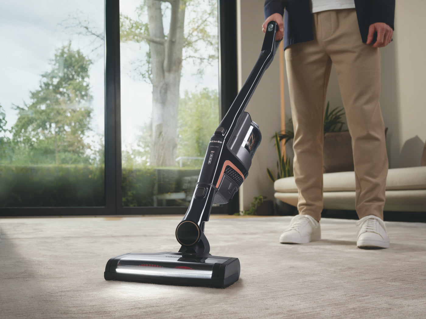 Miele Triflex HX2 Cat & Dog cordless stick vacuum in use on a hardwood floor in a bright modern room.