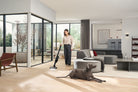 Woman vacuuming a hard floor with the Miele Triflex HX3 Cat & Dog in Obsidian Black while a dog rests nearby.